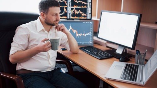 Young businessman is sitting in office at table, working on computer with many monitors,diagrams on monitor. Stock broker analyzes binary options charts.Hipster man drinking coffee,studying
