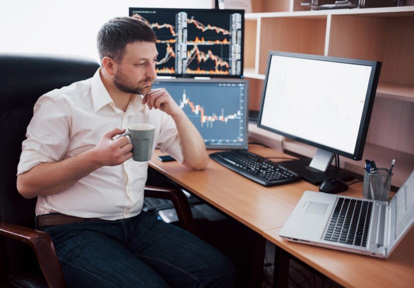 Young businessman is sitting in office at table, working on computer with many monitors,diagrams on monitor. Stock broker analyzes binary options charts.Hipster man drinking coffee,studying