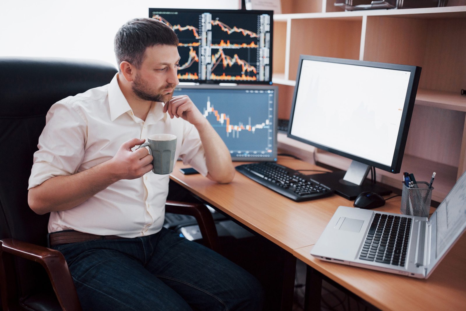 Young businessman is sitting in office at table, working on computer with many monitors,diagrams on monitor. Stock broker analyzes binary options charts.Hipster man drinking coffee,studying